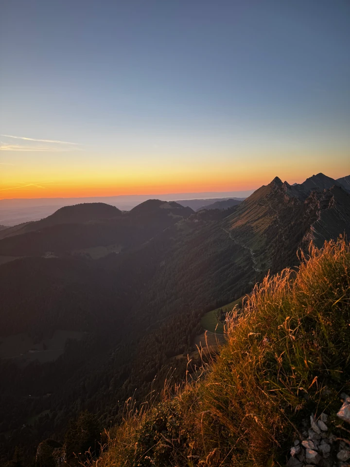 Succession de crêtes montagneuses au coucher du soleil, avec des herbes éclairées au premier plan.